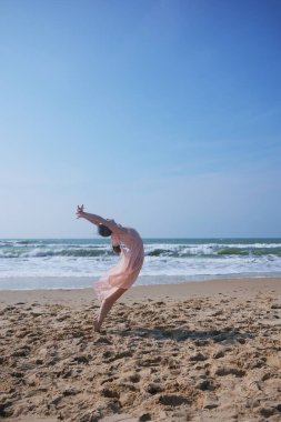 Açık pembe elbiseli çıplak ayaklı bir bayan dansçı, güneşli bir plajda dramatik bir geri dönüş yapar. Zarif hareket, arkasındaki okyanus dalgalarıyla çelişiyor.. 