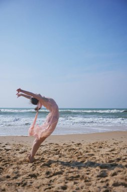 Yumuşak pembe elbiseli bir bayan dansçı kumlu bir plajda anlamlı bir sırt kemeri sergiliyor. Arkasından dalgalar çarpıyor, parlak mavi gökyüzünün altında yalınayak yürürken..