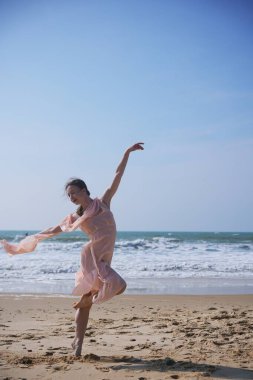 Açık pembe şifon elbiseli zarif bir kadın parlak, kumlu bir plajda zarif bir bale pozu veriyor. Okyanus dalgaları, berrak mavi bir gökyüzünün altında, dinginliği çağrıştırıyor.. 