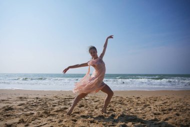 Açık pembe şifon elbiseli bir bayan dansçı deniz kenarında çıplak ayakla kumda zarifçe hareket eder. Anlamlı duruşu zarafeti, özgürlüğü ve doğal hareketi yansıtıyor.