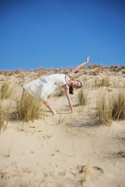 Beyaz elbiseli çıplak ayaklı bir kadın altın kum tepelerinde dinamik ve zarif bir poz veriyor. Mavi gökyüzü ve kuru otlar barışçıl, doğal ve sanatsal bir atmosfer yaratır..