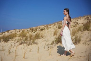 Sırtsız beyaz elbiseli genç bir kadın sıcak güneş ışığıyla açık mavi bir gökyüzünün altında kuru bir kum tepesinde yalınayak yürüyor.. 