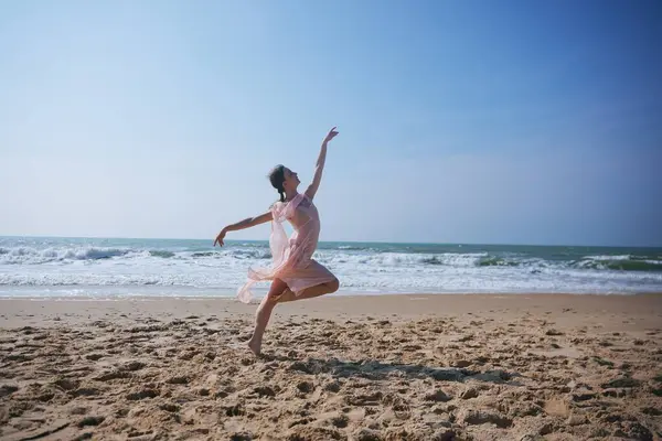 Açık pembe şifon elbiseli zarif bir kadın güneşli bir sahilde yalınayak zıplıyor. Geniş kolları ve rüzgarın savurduğu kumaşı güçlü bir zarafet ve hareket anı yaratır..