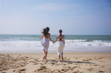 Güneşli bir günde el ele tutuşan ve okyanus kıyısında oynayan iki neşeli kadın, özgürlük ve arkadaşlığın tadını çıkarıyorlar..