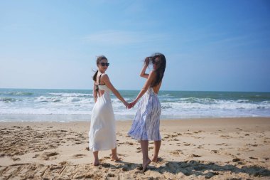 Yazlık elbiseler içinde iki şık kadın el ele tutuşmuş ve deniz kenarında gülümsüyor. Okyanus kenarında güneşli bir günün tadını çıkarıyorlar..