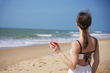 Bir kadının kokteyl bardağını tutarken, sahilde dikilirken ve güneşli bir günde okyanus manzarasının keyfini çıkarırken.. 