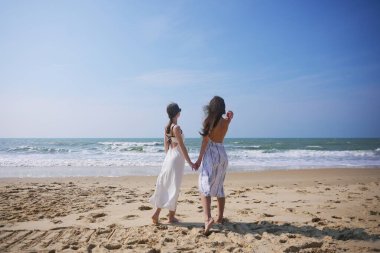Yazlık elbiseli iki kadın sahilde çıplak ayakla yürürken el ele tutuşup güneşli ve esintili bir deniz gününün tadını çıkarıyorlar.. 