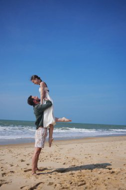 Genç bir çift arasında romantik bir an. Erkek, kadını neşeyle güneşli ve huzurlu bir sahilde kaldırır. Mükemmel bir yaz tatili sırasında kaygısız ve sevgi dolu bir bağı paylaşıyorlar.. 