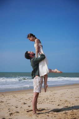 Genç ve neşeli bir çift deniz kenarında romantik bir anı paylaşırken erkek kadını neşeyle kaldırıyor. Açık mavi gökyüzü, yumuşak dalgalar ve altın kum aşk ile mükemmel bir yaz ortamı yaratır.,