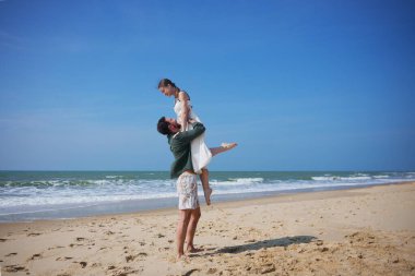 Genç adam gülümseyen kadını dalgalar ve mavi gökyüzüyle kumlu sahilde kucaklıyor. Bu romantik ve eğlenceli an kaygısız yaz titreşimlerini, aşkı ve mutluluğu yakalar.