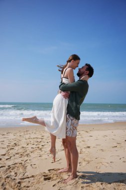 Sevgi dolu bir çift, güneşli bir plajda sıcak bir kucaklaşmayı paylaşır. Erkek, kadını gülümserken kaldırır. Okyanus dalgaları ve parlak gökyüzü ile çevrili bu an neşeyi yakalar..