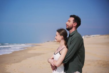 Romantik çift, okyanus esintisi ve sakin sahil manzarasıyla geniş ve boş bir plajda huzurlu bir kucaklaşmanın tadını çıkarıyor..