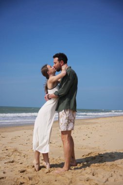 Genç bir çift, okyanus dalgaları ve altın kumlarla çevrili güneşli bir sahilde şefkatli bir öpücüğü paylaşıyor. Bu romantik sahne derin duygusal bağlar kurar.
