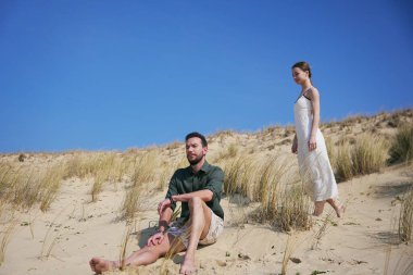 Genç adam güneşli kumullarda huzur içinde otururken, beyaz elbiseli bir kadın açık mavi gökyüzünün altında yalınayak yürüyor.. 