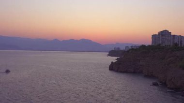 Kıyı uçurumları ve şehir silueti üzerinde akşam alacakaranlığı, uzakta dağ silueti ile sakin deniz, gün batımından sonra okyanus ufkunun huzurlu panoramik manzarası yumuşak pastel gökyüzü
