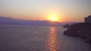 Kıyı uçurumları ve şehir silueti üzerinde akşam alacakaranlığı, uzakta dağ silueti ile sakin deniz, gün batımından sonra okyanus ufkunun huzurlu panoramik manzarası yumuşak pastel gökyüzü