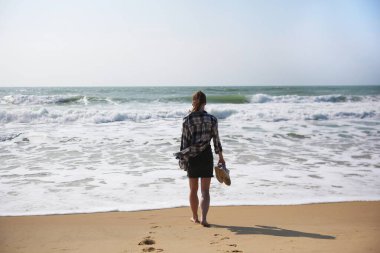 Okyanus dalgalarına doğru kumsalda yürüyen, elinde ayakkabıları olan, sakin deniz melteminin tadını çıkaran bir kadının arka görüntüsü..