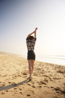 Çıplak ayakla kumsalda yürüyen, kolları özgürce yükselen, güneş ışığı ve okyanus esintisinin tadını çıkaran bir kadının arkası.. 