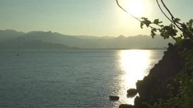 Kayalık kıyı şeridi ile sakin bir günbatımı sahnesi, uzak sıradağlar, ve bereketli yeşil ağaçlar, sakin ve manzaralı temalar için ideal..