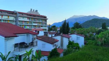 Renkli bir otelin insansız hava aracı fotoğrafı ve Antalya 'da yemyeşil ağaçları, palmiyeli sokakları ve uzak dağları olan yerel evler.. 