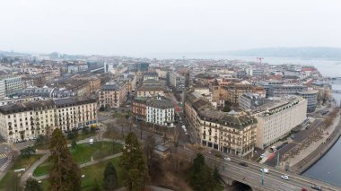 Cenevre, İsviçre 04.11.2025 Geleneksel binalar, dolambaçlı sokaklar, nehir kenarındaki bir gezinti alanı ve bulutlu gökyüzünün altındaki uzak gölü gösteren insansız hava aracı görüntüsü. 
