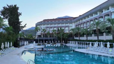 Antalya, Türkiye 04.11.2025 Çağdaş Otel geniş bir yüzme havuzu, palmiye ağaçları ve geniş açılı perspektiften yumuşak akşam ışığı altında çekilen güneş pansiyonları. 