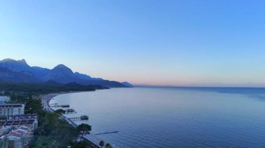 İHA görüntüsü, yumuşak akşam ışığında oteller, sahil ve görkemli dağ sıraları olan bir sahil beldesine iniyor..