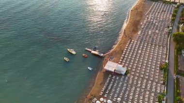 Sakin deniz, güneş yansıması, plaj şezlongları, rıhtımlar ve sıcak ışık altındaki beyaz şehir binalarının hava günbatımı görüntüleri..