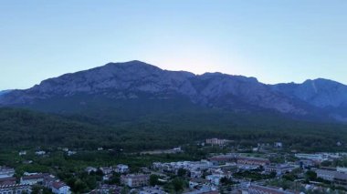 Akşam drone görüntüleri, sık ormanla görkemli dağ sırasının arasına yerleşmiş bir tatil köyünün manzaralı kıyı şeridine doğru yavaşça ilerlediğini gösteriyor..