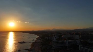 İnsansız hava aracı gün batımında bir plaj beldesine iniyor. Parlayan bir deniz, aydınlatılmış otel binaları ve sıcak altın ışığın altındaki uzak dağları gözler önüne seriyor.