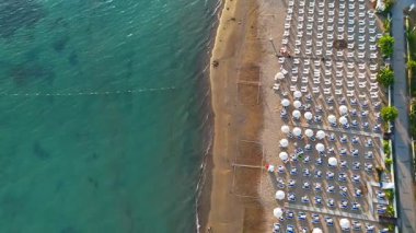 İnsansız hava aracı, geniş gölgeli dinlenme alanları ve sakin denize doğru uzanan geniş bir rıhtımın bulunduğu bir sahil üzerinde çaprazlama uçar ve rahat bir sahil manzarası yaratır.