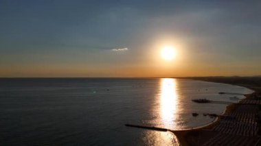 Gün batımında sakin bir kıyı şeridinde yükselen bir İHA 'nın hava görüntüleri, sakin su, rıhtım, tekne ve altın bir ufuk üzerindeki güneş yansımalarını yakalıyor..