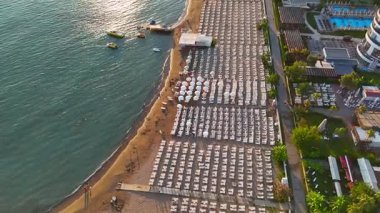 Düzenli güneş şezlongları ve şemsiyeleri, turkuaz suyu, yüzücüler, botlar ve dengeli bir kıyı sahnesi oluşturan rıhtımların olduğu kumlu bir sahilin tepeden aşağı bakan drone görüntüsü..