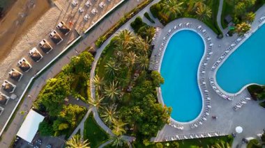 Gün batımında tropikal bir tatil köyünün tepeden aşağı drone görüntüsü, plaj kabinleri, güneş şezlongları ve palmiye ağaçlarıyla çevrili lüks bir havuz..