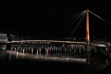 Lyon 'daki Passerelle du Palais de Justice' in gece manzarası, sakin suyun üzerinde sıcak ışıklarla aydınlatılmış modern bir yaya asma köprüsü..