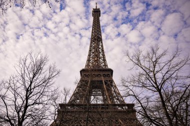 Eyfel Kulesi, Paris 'teki bulutlu mavi gökyüzünün altında çıplak kış ağaçları arasında yükseliyor. Seyahat, mimari ve mevsimsel temalar için yatay manzara görünümü.