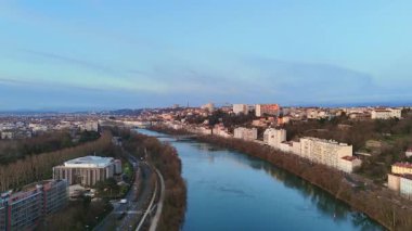 Fransa 'nın Lyon kentinden akan bir nehrin manzaralı perspektifi, sakin mavi bir sabah gökyüzü altında konut binaları ve yeşil alanlar..