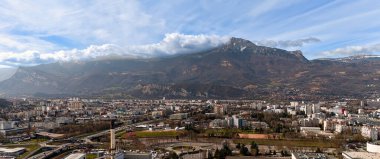Yüksek dağlarla çevrili bir şehrin insansız hava aracı görüntüsü ön planda yükselen bir baca ve gökyüzünde sürüklenen dramatik bulutlar.