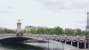 Paris, Fransa 06.07.2025 Süslü Pont Alexandre III köprüsünün sokak lambalarıyla panoramik görüntüsü, insanlar karşıya geçiyor, ve Eiffel Kulesi bulutlu bir günde arka planda.