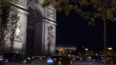 Arabalar geceleri Paris 'teki aydınlık Zafer Takı' nın yanından geçerken, kasvetli bir şehir manzarasında ikonik mimari ve dinamik şehir hareketi sergiliyorlar..