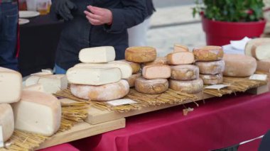 Satıcıların arka planda örnekleri dilimlediği, saman kaplı bir tezgahta sergilenen çeşitli tekerlek ve zanaat peyniri parçaları..