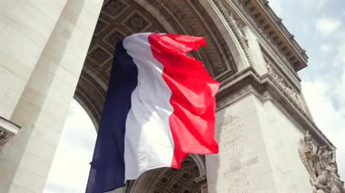 Paris 'teki Zafer Takı' nın altında rüzgarda dalgalanan canlı Fransız üç renkli bayrağı. Detaylı taş mimarisi ve arka planda mavi gökyüzü..