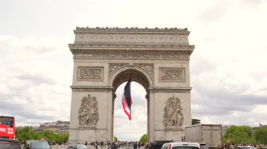 Paris, Fransa 06.08.2025 Zafer Takı Anıtı manzarası turistler ve trafik bulutlu bir öğleden sonra kavşağı doldururken Fransız ulusal bayrağı kemerin altında dalgalanıyor..