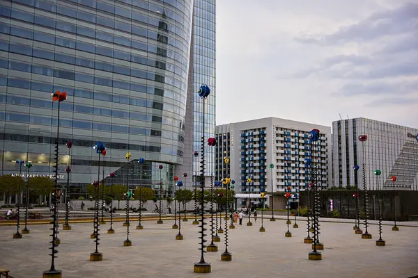 La Defense 'da renkli şehir heykelleri duruyor. Etrafı cam kulelerle çevrili modern mimari ve bulutlu bir gökyüzünün altında yürüyen insanlar.
