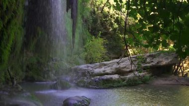 Bir şelalenin arkasından, yosunlu kayaların ve orman akıntısının üzerinden ön planda damlacıklar ve yapraklı Türk zeminini aydınlatan güneş ışığının olduğu bir manzaraya bakın..