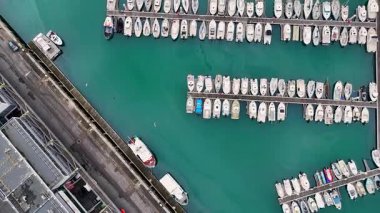 Üst taraftaki drone görüntüleri paralel ahşap rıhtımlar boyunca yanaşmış tekneleri gösteriyor. Durgun turkuaz su, temiz geometrik yerleşim ve görünür yat siluetleri var. 