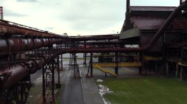 Eski bir endüstriyel kompleksteki zemin katlı yürüyüş yolları ve paslı boru hatları modernize edilmiş kamusal alanda korunmuş çelik yapıları gösteriyor.