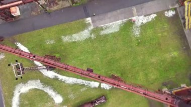 Eski bir fabrika binasının yanından geçen paslı endüstriyel boru hatlarının hava drone görüntüsü, geometrik bir düzendeki çim ve asfalt ile çelişiyor. 