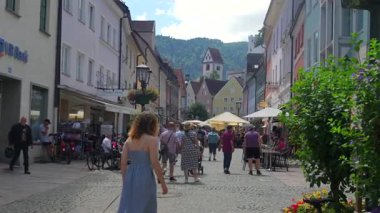 24 Temmuz 2025 'te Fuessen Almanya' da kaldırımlı bir cadde boyunca yürüyen turistler, tarihi binalar ve yol boyunca uzanan hareketli kafe teraslarıyla 