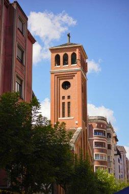 Açık mavi gökyüzünün altındaki binalar ve ağaçlar tarafından çerçevelenmiş kırmızı tuğlalı bir kilise kulesinin dikey görüntüsü